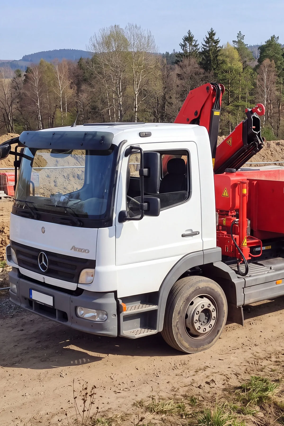 Bílý nákladní vůz Mercedes-Benz Atego s hákovým nosičem a hydraulickou rukou na staveništi