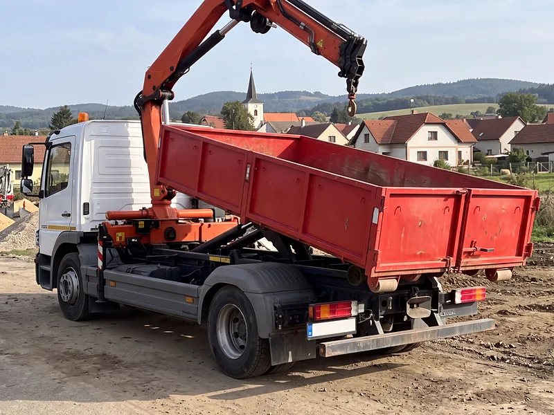 Nákladní vůz Mercedes-Benz Atego zezadu se sklopeným hákovým kontejnerem na kraji obce